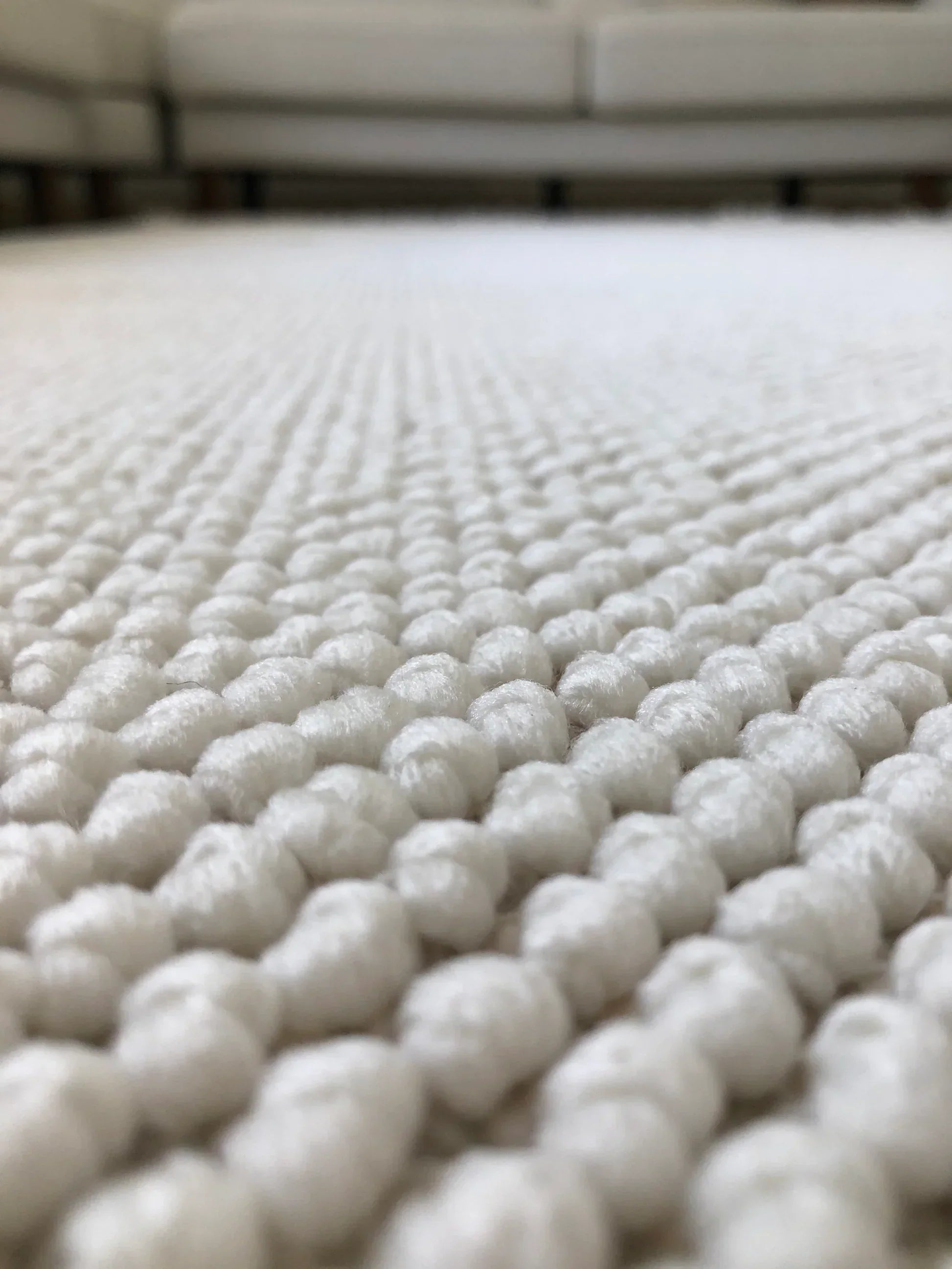 Beyaz renkli, dokulu, kalın örgülü halı yüzeyi, arka planda açık renkli koltuklar