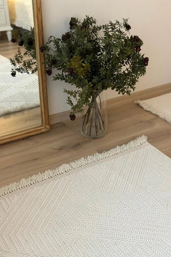 Ahşap çerçeveli ayna yanında cam vazoda yapay yeşil ve koyu kırmızı çiçekler, açık renk dokuma halı