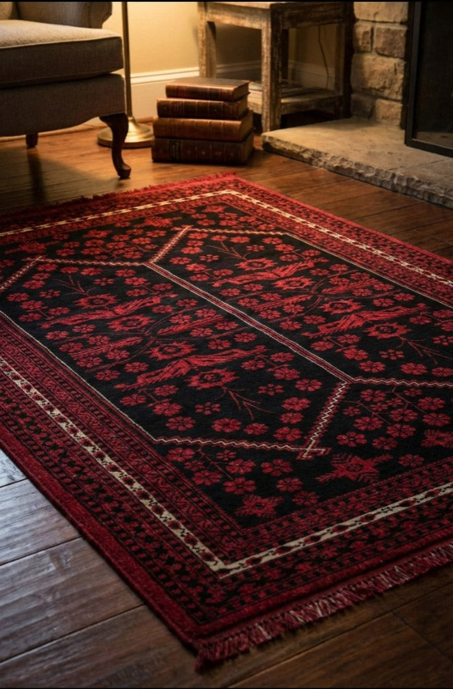 Klasik kırmızı ve siyah motifli Afgan halısı, ahşap zemin ve taş şöminenin önünde, yanındaki koltuk ve kitaplarla