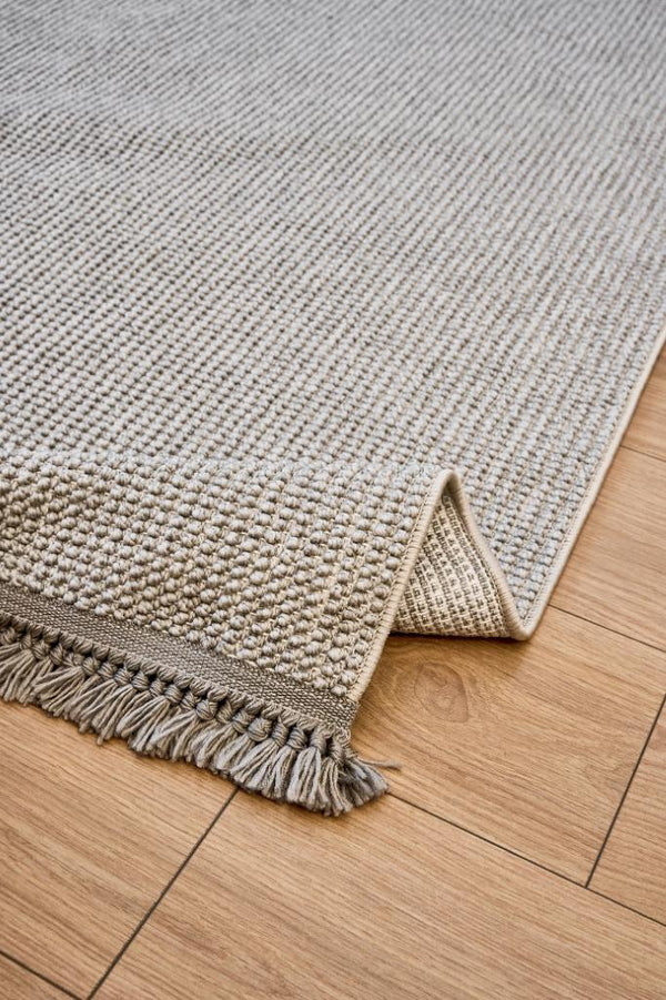 Gri renkli dokuma makine halısı, ahşap zemin üzerinde kıvrılmış ve püsküllü kenar detaylı