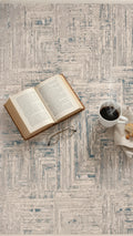 Mavi bej desenli halı üzerinde açık kitap, metal çerçeveli gözlük ve buharlı kahve fincanı